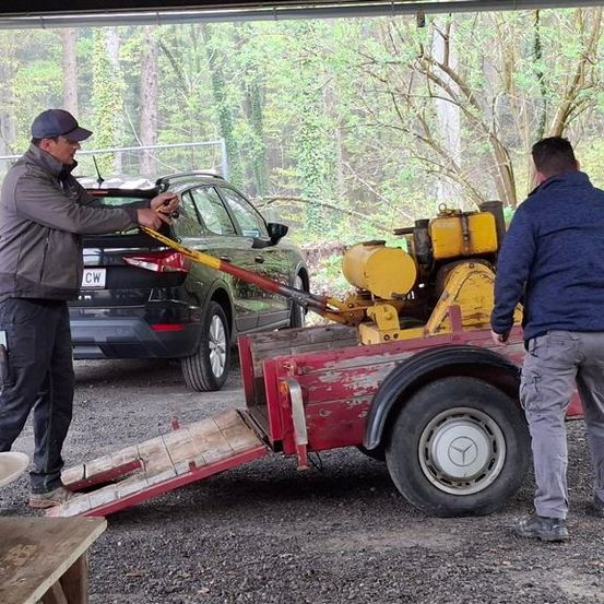 Zwei Männer laden einen schwarzen SUV auf einen roten Anhänger mit gelben Maschinen. Ein Mann hält das Auto, während der andere den Anhänger stützt. Sie befinden sich in einer Garage mit Kies und Bäumen.