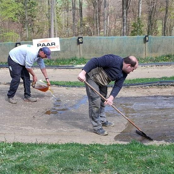 Zwei Männer arbeiten auf einer nassen Straße mit den Nummern 10 und 9 an einer Wand. Einer gießt Flüssigkeit ein, während der andere fegt. Bäume und ein Zaun sind im Hintergrund.
