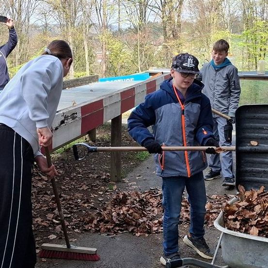 Menschen räumen Blätter vom Boden auf, wobei eine Frau einen Besen benutzt, ein Junge einen Rechen hält und ein anderer Junge einen mit Blättern gefüllten Schubkarren schiebt. Eine andere Person steht hinter ihnen.