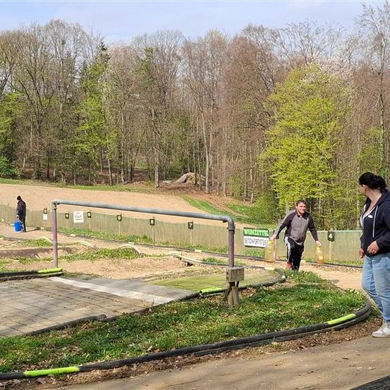 Mehrere Personen befinden sich auf einer Erdbahn in einer Outdoor-Sportanlage. Eine Person beugt sich vor und zwei Personen stehen in der Nähe der Bahn. Bäume umgeben das Gebiet.