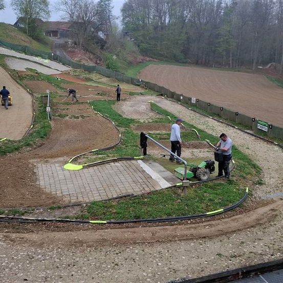 Mehrere Personen arbeiten auf einer Erdbahn, eine mit einem Traktor, umgeben von grünem Gras und einem eingezäunten Feld. Bäume und Gebäude sind im Hintergrund.