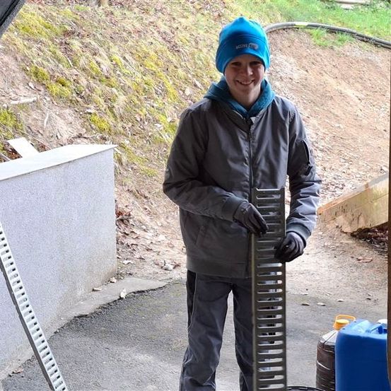 Eine Person steht in einem Außenbereich, hält ein Metallobjekt und scheint für ein Foto zu posieren. Eine Wand ist links, und eine Stahlleiter ist an der Wand befestigt. Ein Stahlstab ist auf dem Boden befestigt, und eine Tafel ist an der Wand befestigt. Hinter der Person befindet sich ein Grasfeld.