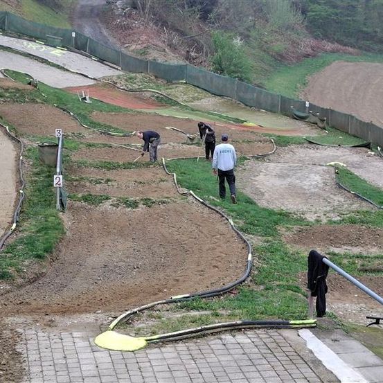 Eine Draufsicht auf eine Erdrennstrecke mit Menschen, die daran arbeiten. Drei Personen benutzen Schaufeln, um Erde zu bewegen, während eine weitere Person bei einem gelben Objekt steht. Die Strecke ist von einem Zaun umgeben, und dahinter befindet sich ein Feld.