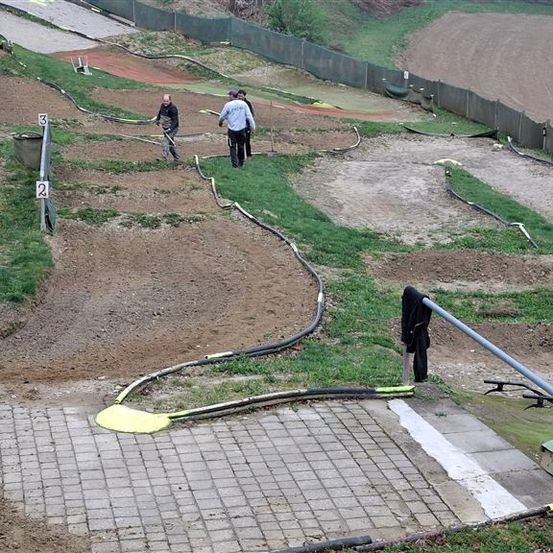 Eine Dirt-Rennstrecke wird vorbereitet. Mehrere Personen arbeiten daran. An den Seiten sind Nummern angebracht, die verschiedene Abschnitte kennzeichnen. In der Nähe befindet sich ein grünes Maschinenfahrzeug.