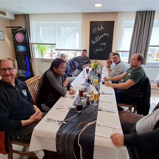 Eine Gruppe von Männern sitzt an einem Tisch in einem Raum mit einer Tafel, Fenstern und Pflanzen. Sie lächeln und scheinen Getränke und Essen zu genießen.