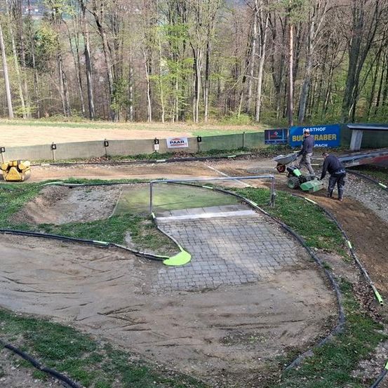 Eine Erdbahn mit drei Arbeitern, die einen Radlader benutzen. Die Strecke ist von einem grünen Zaun umgeben, mit Bannern und Bäumen im Hintergrund.