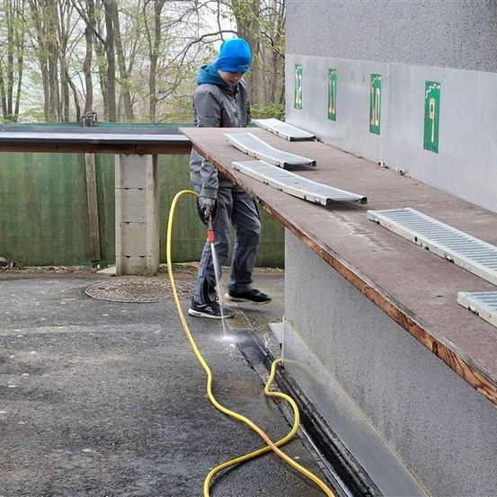 Ein Mann mit blauer Mütze und Turnschuhen reinigt mit einem Schlauch eine Betonwand mit nummerierten Schlitzen.