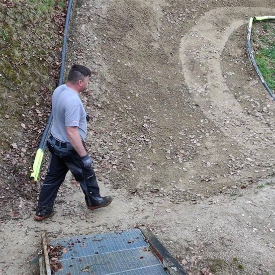 Ein Mann in einem grauen T-Shirt und Hose läuft auf einem steilen Pfad mit Kies und kleinen Steinen. Er hält ein gelbes Werkzeug und ist wahrscheinlich ein Arbeiter.