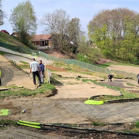 Eine Gruppe von Arbeitern baut eine Dirtbike-Strecke. Sie tragen Schutzausrüstung und verwenden Werkzeuge. Die Strecke ist von einem Zaun und Grün umgeben.