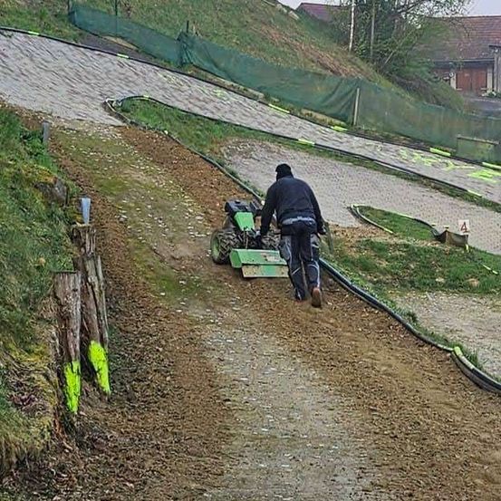 Ein Mann geht einen Pfad neben einem grünen Traktor auf einer Schotterpiste entlang, mit einem grünen Zaun und einem Haus im Hintergrund.
