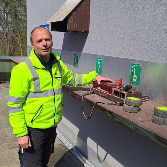 Ein Mann in einer Warnweste steht neben einer Wand mit nummerierten Schildern und zeigt auf eine rote Maschine mit Kabeln und Rädern auf einem Sims.