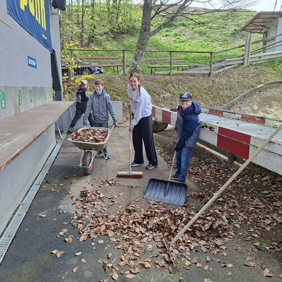 Mehrere Personen räumen Blätter auf einem Weg auf. Eine Person benutzt einen Schubkarren, während andere Besen und Schaufeln haben. Ein Gebäude mit einem blauen Banner befindet sich in der Nähe.