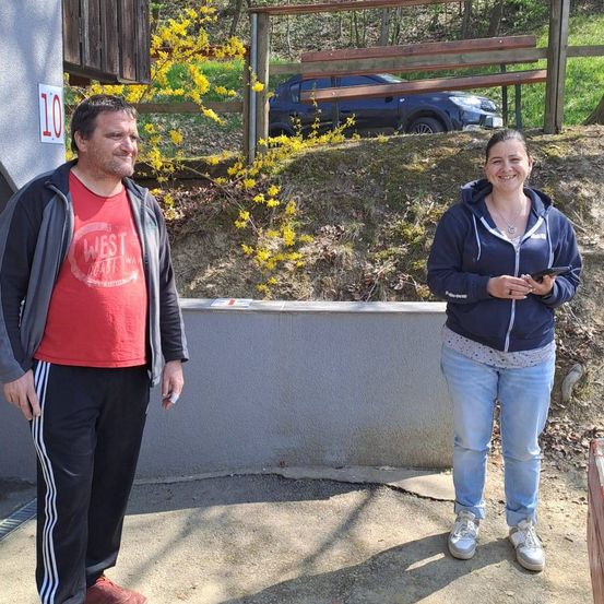Ein Mann und eine Frau stehen vor einer Wand mit einem Schild mit der Nummer 10. Der Mann trägt ein rotes T-Shirt und schwarze Hose. Die Frau hat eine blaue Jacke an und hält ein Telefon. Ein Auto ist hinter einem Zaun geparkt.