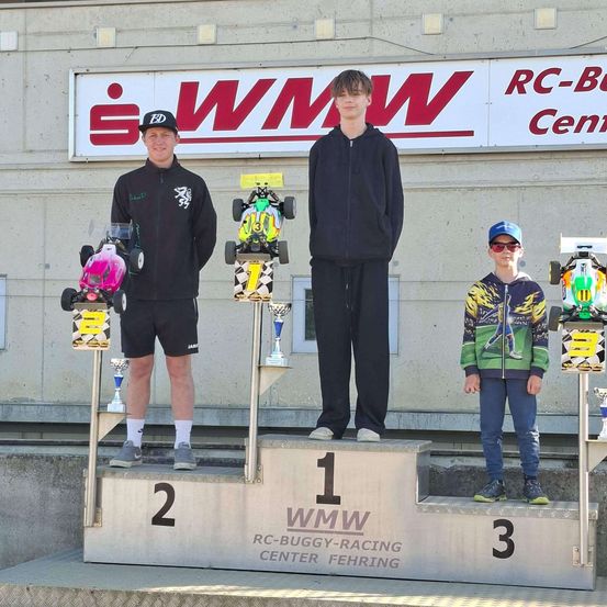 Drei Fahrer stehen auf einem Podest. Zwei halten Pokale, einer in Schwarz, der andere in buntem Design. Der mittlere Fahrer steht zwischen ihnen. Dahinter zeigt ein Gebäude ein Schild mit der Aufschrift 'iWMW RC-Buggy-Racing Center'.