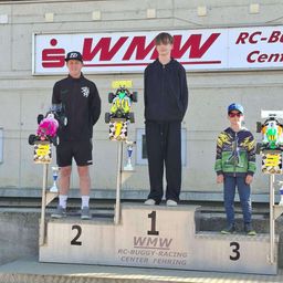 Drei Fahrer stehen auf einem Podest. Zwei halten Pokale, einer in Schwarz, der andere in buntem Design. Der mittlere Fahrer steht zwischen ihnen. Dahinter zeigt ein Gebäude ein Schild mit der Aufschrift 'iWMW RC-Buggy-Racing Center'.