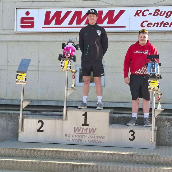 Zwei Männer stehen auf einem Podest mit Trophäen. Einer trägt einen schwarzen Mantel, der andere eine rote Kapuzenjacke. Ein Schild hinter ihnen trägt die Aufschrift 'WMW' und 'RC-Buggy Center'.