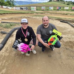 Zwei Männer posieren für ein Foto auf einer Erdrennstrecke, einer mit einer Medaille und beide halten Spielzeugautos.