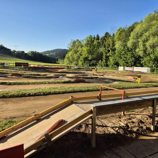 Eine Dirtbike-Strecke mit einer hölzernen Rampe unter einem klaren Himmel, umgeben von üppigem Grün und Bergen in der Ferne.