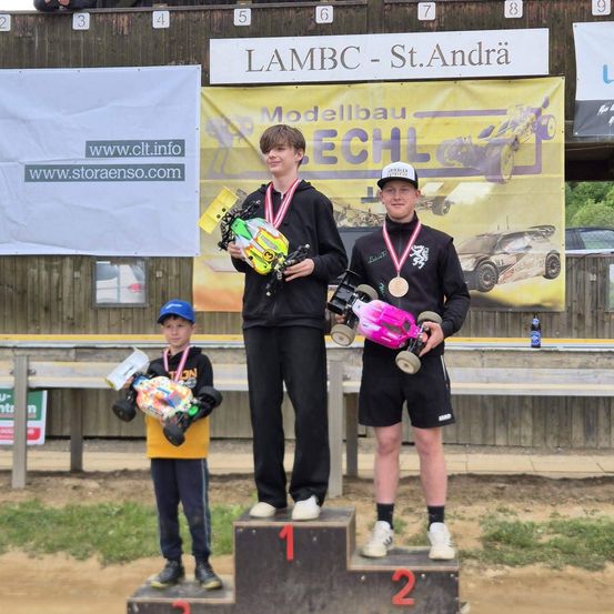 Drei junge Rennfahrer stehen auf einem Podest. Zwei Männer halten Trophäen und Medaillen. Ein Junge steht darunter mit seiner eigenen Trophäe. Hinter ihnen steht ein Banner mit 'LAMBC - St. Andra'.