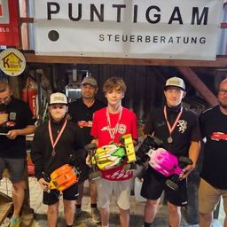 Gruppe von Menschen posiert mit Spielzeugautos und Medaillen vor einem Gebäude mit einem Schild, das Puntigam Steuerberatung liest.