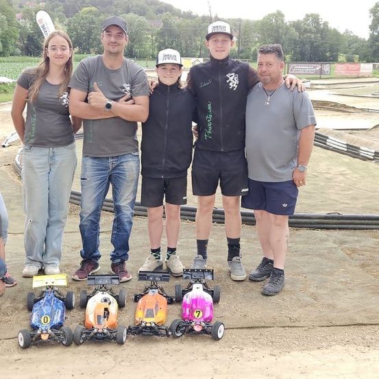 Fünf Personen stehen in einer Gruppe auf einer Erdbahn mit fünf Spielzeugautos mit den Nummern 0, 7 und anderen. Sie tragen Freizeitkleidung. Der Hintergrund ist unscharf, mit sichtbaren Bäumen und Pflanzen.
