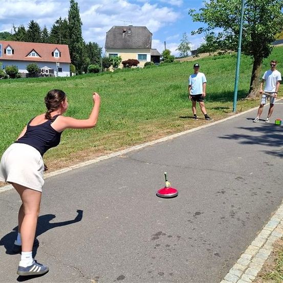 Eine Frau in einem schwarzen Tanktop und einer weißen Shorts wirft einen Ball auf ein rotes Ziel auf der Straße, während zwei Männer in weißen Hemden und schwarzen Shorts in der Nähe stehen.