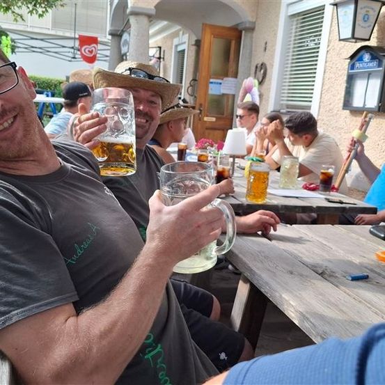Eine Gruppe von Männern sitzt draußen an einem Tisch und hält Biergläser in der Hand. Ein Mann lächelt und hält ein Glas hoch.