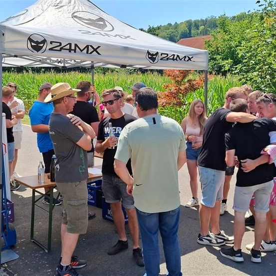 Eine Gruppe von Menschen versammelt sich unter einem Zelt mit dem 24MX-Logo, einige trinken und andere unterhalten sich. In der Nähe steht ein Tisch mit Flaschen und einem Kühler.