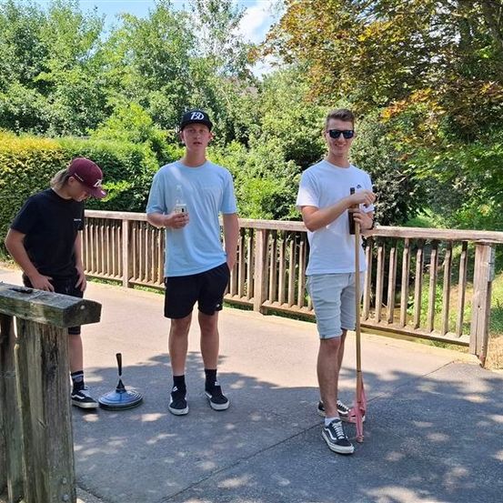 Drei junge Männer stehen auf einem Betonweg, zwei halten Flaschen und einer einen Stock. Sie sind lässig gekleidet und scheinen für ein Foto zu posieren. Hinter ihnen ist ein Holzzaun und Grünpflanzen zu sehen.