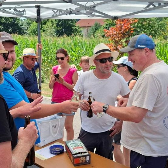Eine Gruppe von Menschen, einige mit Hüten und Sonnenbrillen, versammelt sich unter einem Zelt und stoßen mit Bierflaschen an. Vor ihnen steht ein Tisch mit einer Box und einem Beutel.