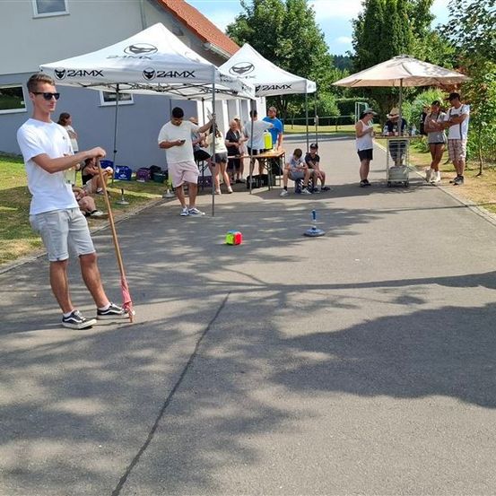Eine Gruppe von Menschen ist in einem Außenbereich versammelt, möglicherweise für ein Spiel, mit aufgestellten Zelten und einem Tisch. Ein Mann in einem weißen Shirt hält einen Stock und schaut auf einen Ball.