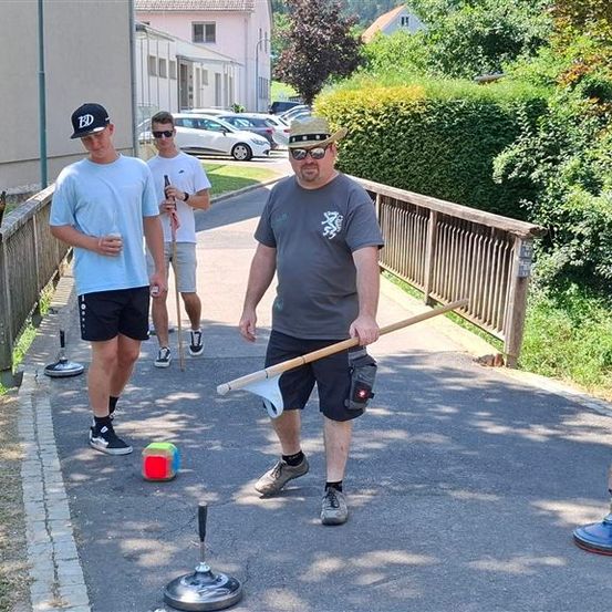 Drei Männer spielen ein Spiel auf einem gepflasterten Weg. Ein Mann hält einen Stock und eine Flagge, während die anderen beiden zusehen.