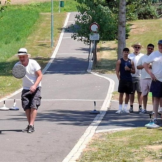 Ein Mann wirft einen Frisbee auf einem Pfad, während andere zuschauen. Sie sind an einem sonnigen Tag, mit Gras und Bäumen in der Nähe.