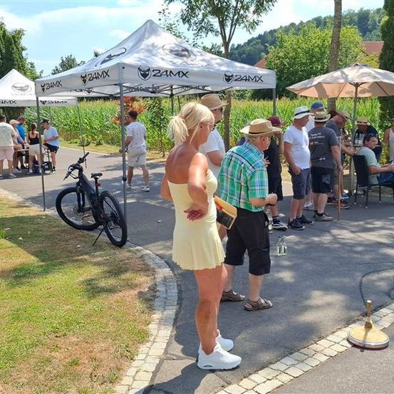 Eine Gruppe von Menschen versammelt sich um ein Zelt und Schirme, eine Frau in einem gelben Kleid steht neben einem Fahrrad. Ein Mann mit Hut hält ein Buch.