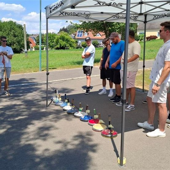 Eine Gruppe von Männern steht unter einem Zelt, möglicherweise in Vorbereitung auf ein Spiel. Ein Mann hält einen Stock, und mehrere bunte Objekte sind auf dem Boden aufgereiht.