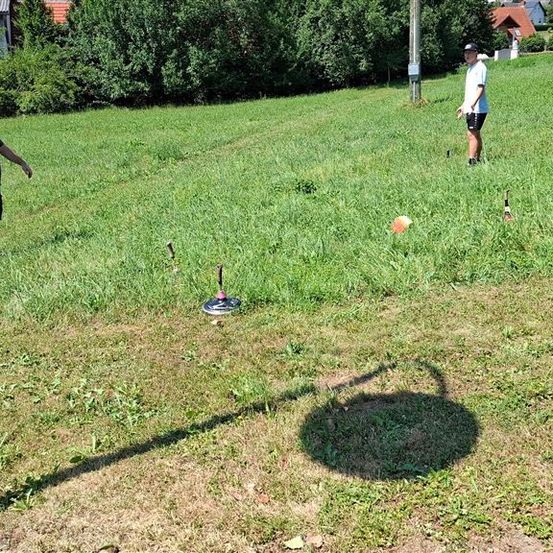 Zwei Männer spielen ein Cornhole-Spiel auf einem Grasfeld mit mehreren Zielen und verstreuten Bällen.