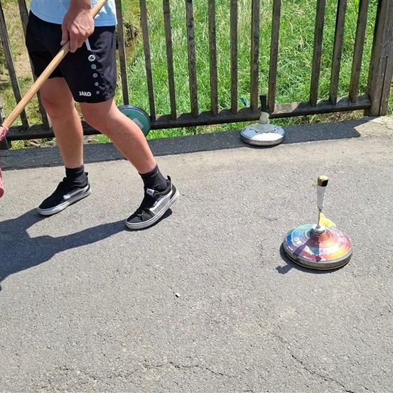 Eine Person in schwarzen Shorts und Turnschuhen hält einen Stock mit einem rosa Band und steht auf Beton in der Nähe eines Holzzauns.