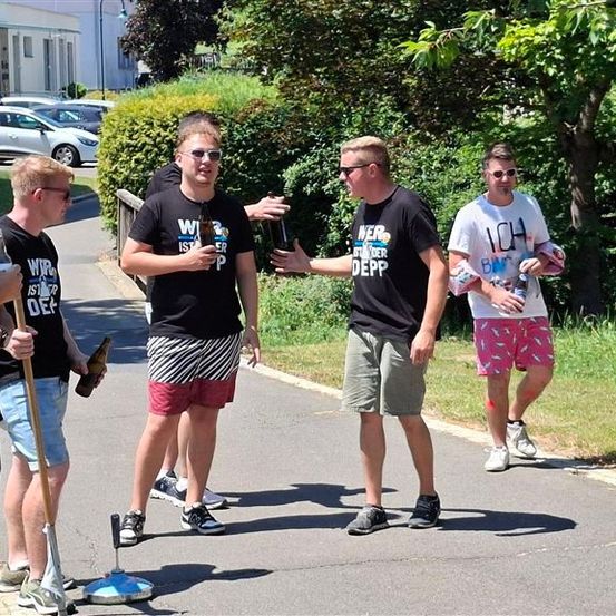 Fünf Männer mit Sonnenbrillen und Flaschen in der Hand gehen auf einem Bürgersteig. Ein Mann hält einen Besen.