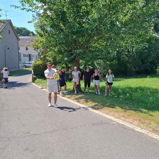 Eine Gruppe von Menschen steht auf einem Bürgersteig, einige halten Frisbees, unter einem großen Baum an einem sonnigen Tag.