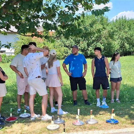 Eine Gruppe von Menschen steht auf einem Feld, einige tragen Hüte und Turnschuhe, und ein Mann berührt seinen Kopf. Sie sind von Frisbees auf dem Boden umgeben.