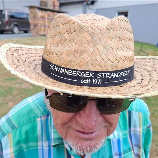 Ein älterer Mann trägt einen Strohhut mit der Aufschrift Schwanberger Strandfest seit 1971. Er trägt eine Sonnenbrille und ein kariertes Hemd.