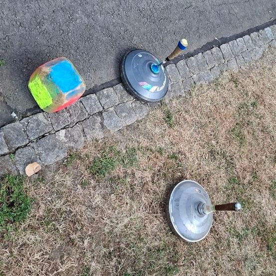 Ein bunter Würfel und zwei Metallobjekte liegen am Boden in der Nähe eines Ziegelstein-Gehwegs. Der Würfel hat blaue, grüne und rote Farbe. Die Metallobjekte ähneln einer Spielzeug-Spitze.