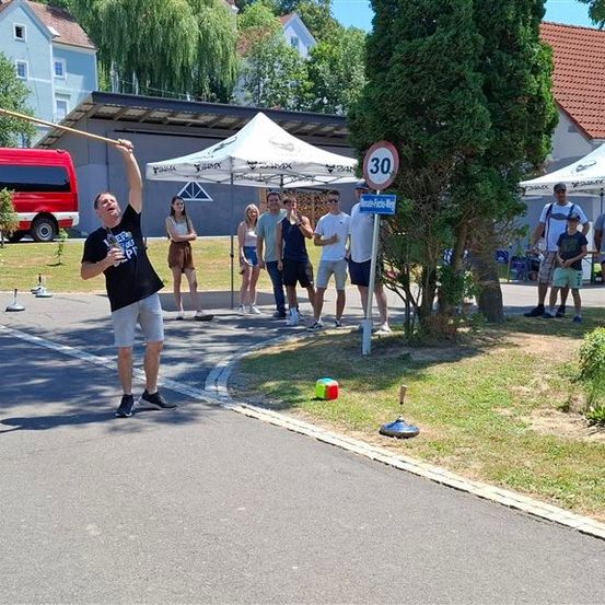 Ein Mann wirft einen Speer, während eine Gruppe von Menschen zusieht, mit Zelten und einem Geschwindigkeitsbegrenzungsschild im Hintergrund.