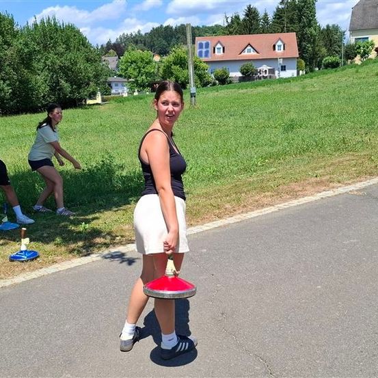 Eine Frau hält ein rotes und silbernes Objekt in der Hand, während sie auf einer Straße geht, mit drei Personen im Hintergrund, die ein Spiel mit blauen Objekten auf einem Grasfeld spielen.