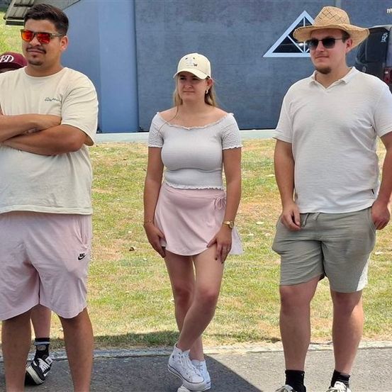 Fünf Personen stehen auf einem Bürgersteig, drei Männer und zwei Frauen. Die Männer tragen Hüte und Sonnenbrillen, und die Frauen tragen ein weißes Hemd und einen rosa Rock. Sie alle schauen in die gleiche Richtung. Hinter ihnen befindet sich ein Gebäude mit einem dreieckigen Design an der Wand.