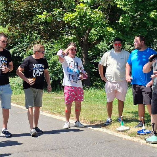 Eine Gruppe von Männern in lässiger Kleidung steht auf einem Pfad, einige halten Flaschen, mit einem Grasbereich und Bäumen im Hintergrund.