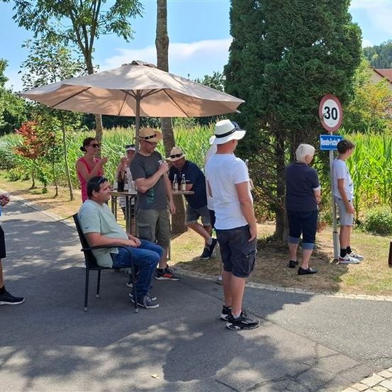 Eine Gruppe von Menschen steht und sitzt um einen Tisch unter einem Schirm, mit einem Geschwindigkeitsbegrenzungsschild und einem Haus im Hintergrund.