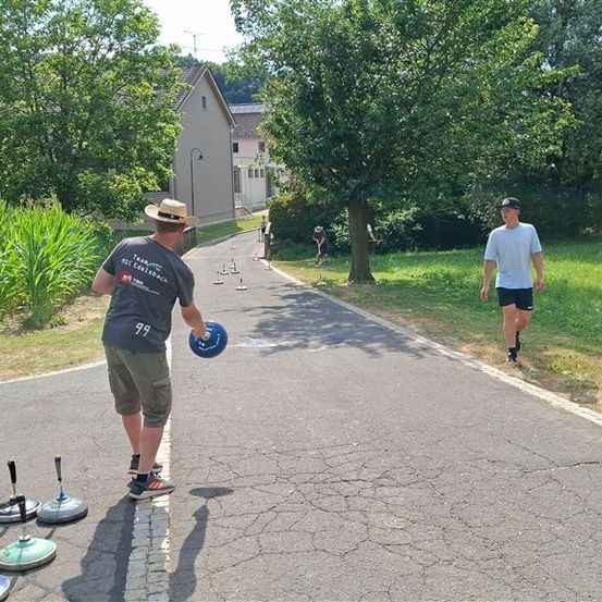 Eine Gruppe von Menschen spielt eine Partie Boccia auf einem gepflasterten Weg in einem Park. Ein Mann wirft einen Ball, während andere zuschauen.