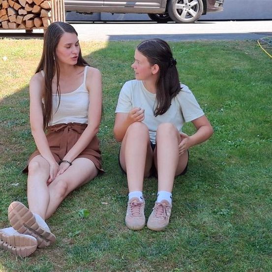 Zwei Mädchen sitzen auf dem Rasen, eines trägt ein weißes Tanktop und einen braunen Rock, das andere ein weißes T-Shirt und Turnschuhe. Sie schauen sich an und scheinen zu sprechen. Dahinter befindet sich eine Holzkonstruktion mit einem Holzstapel obenauf.
