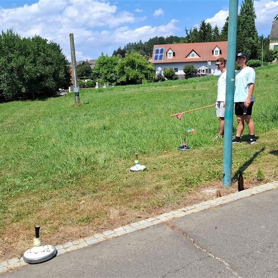 Zwei Männer stehen auf einem Grasfeld mit einem Pfosten und einem Frisbee im Vordergrund. Im Hintergrund befinden sich Häuser und Bäume.
